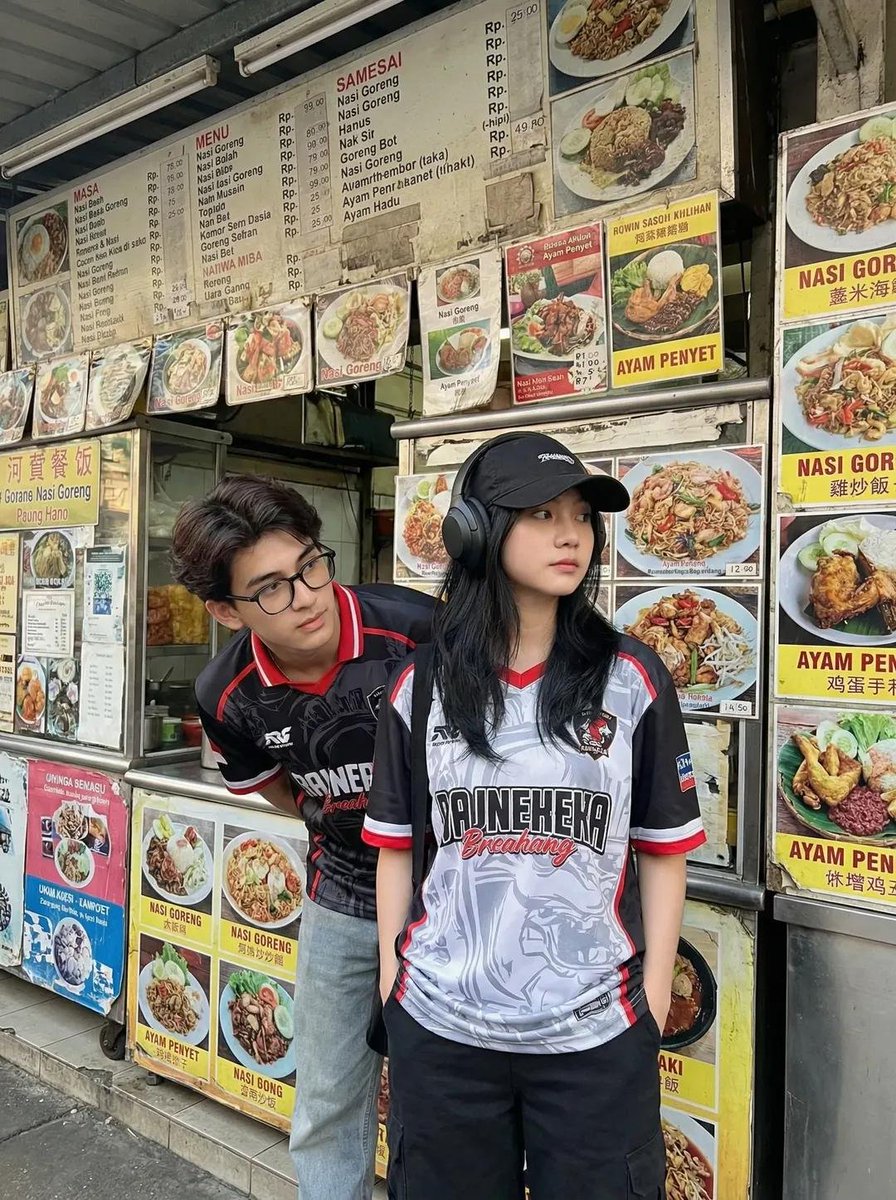 一张抓拍风格的街拍照片:两位年轻的亚洲成年人站在一家小型东南亚街边小吃摊前。小吃摊上挂满了色彩鲜艳的菜单板和食物照片,主打菜品包括 nasi goreng(印尼炒饭)和 ayam…