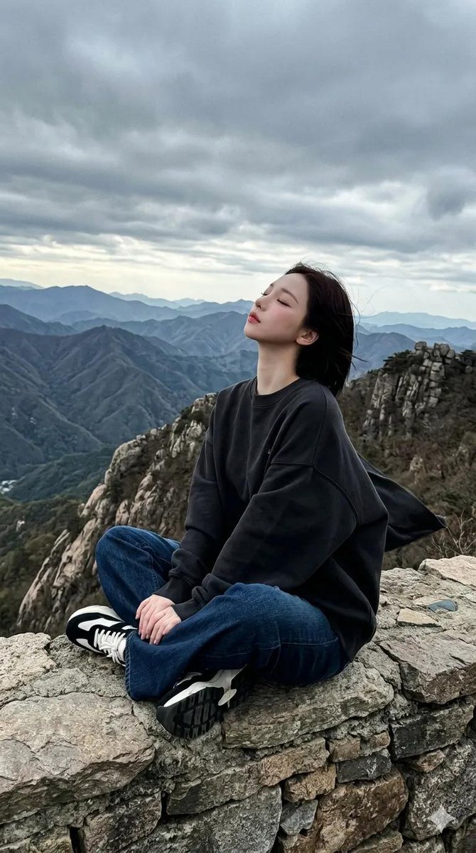 一段电影般的全景画面在戏剧性阴云笼罩的天空下，于一处嶙峋的山脊上徐徐展开。一位女性盘腿坐于石质岩架之上，身着超大号白色印花卫衣、深色阔腿牛仔裤以及黑白配色的厚…