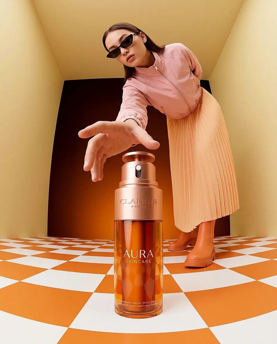 “超现实时尚产品摄影：一只巨大的琥珀色护肤品瓶（Clarins Aura）放置在一个极简盒子房间内的橙白格子地板上，温暖的纯橙色灯光，一位时尚女性戴着太阳镜、…