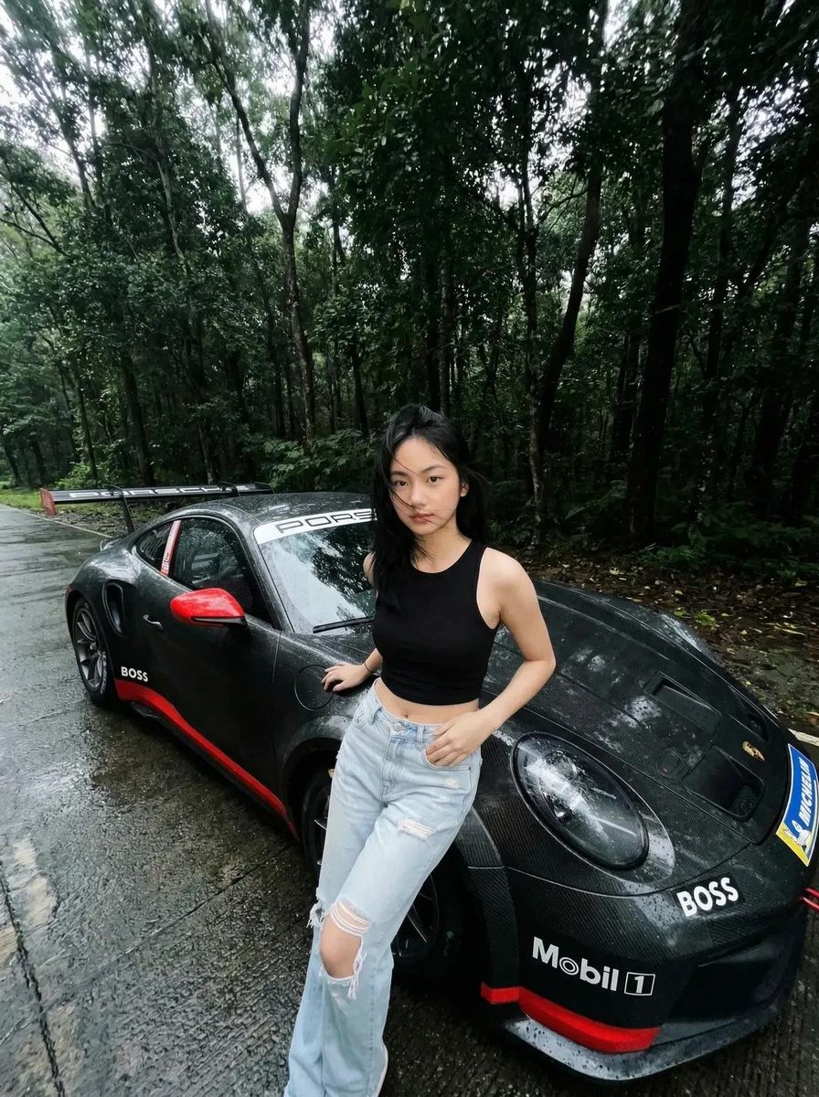一张电影感、超写实的照片：一位年轻亚洲女性站在雨后湿润的森林道路上，旁边是一辆黑色保时捷跑车。她身穿黑色无袖短款上衣和浅蓝色破洞牛仔裤。她的头发略显凌乱，自然飘动，表情平静自信…