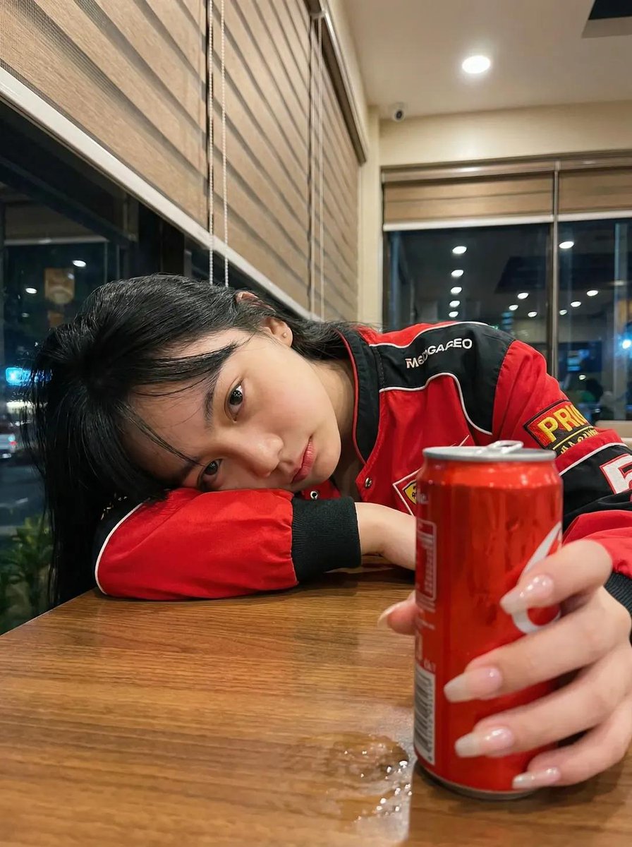 一张超写实的照片：一位年轻女性深夜坐在一间温馨的咖啡馆里，侧着头靠在自己放在木质桌面上的手臂上。她穿着一件红黑配色的赛车风格夹克。表情平静、略带疲惫、若有所思，直视镜头。一只手在…