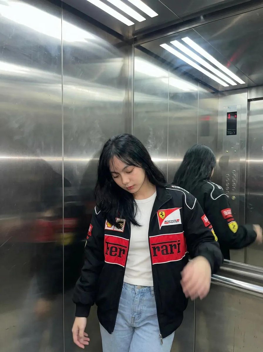 一张超写实的照片：一位年轻女性深夜坐在一间温馨的咖啡馆里，侧着头靠在自己放在木质桌面上的手臂上。她穿着一件红黑配色的赛车风格夹克。表情平静、略带疲惫、若有所思，直视镜头。一只手在…