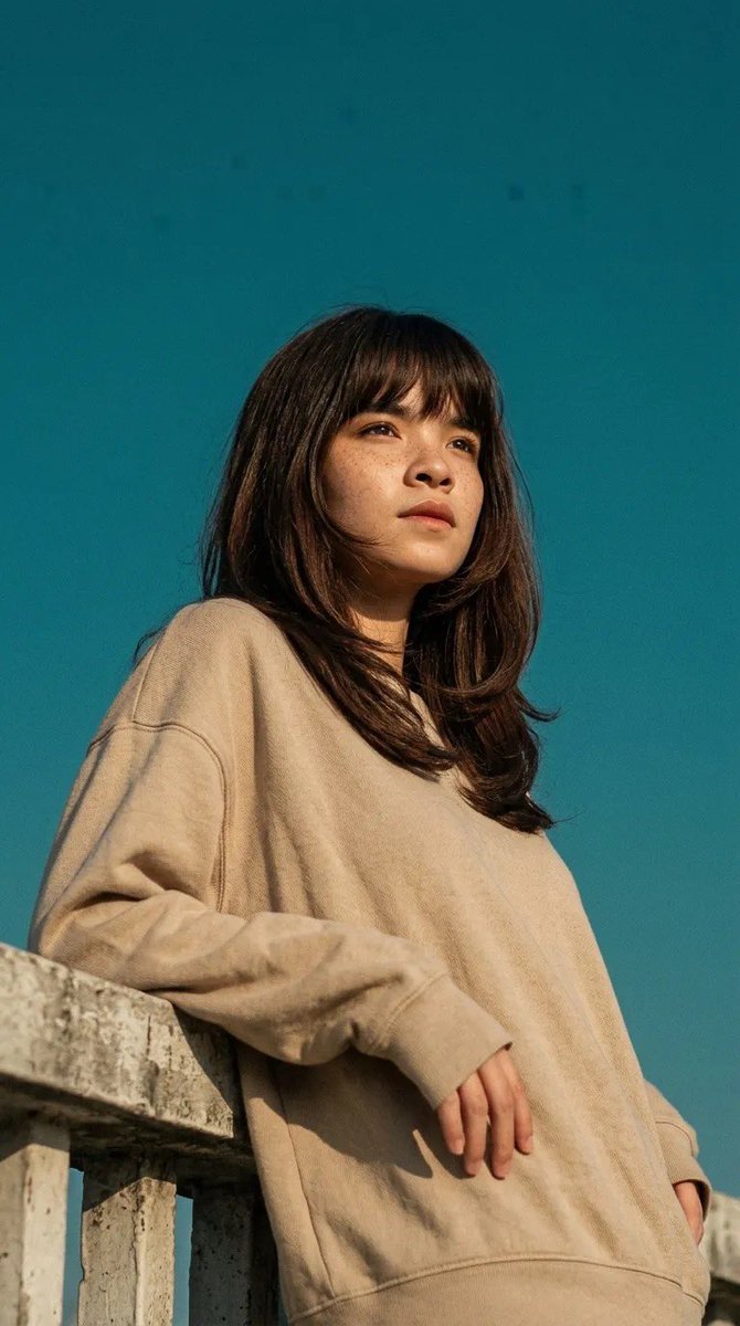 电影感人像照：一位年轻女子倚靠在混凝土栏杆上，身穿宽松米色卫衣，柔和的自然阳光在她脸上投下温暖的色调，背景为清澈的青蓝色天空，淡淡的雀斑，中长深棕色头发配刘海在微风中轻轻飘动，低…