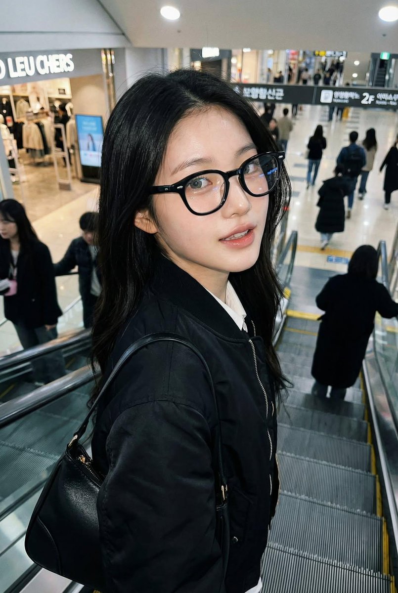 一张像是用 iPhone 拍摄的真实感抓拍照片，画面中是一位女士站在自动扶梯上，面部和身份与参考图完全一致。她戴着黑色玳瑁框眼镜，穿着黑色飞行员夹克，背着一只黑色手提包。姿态自然…