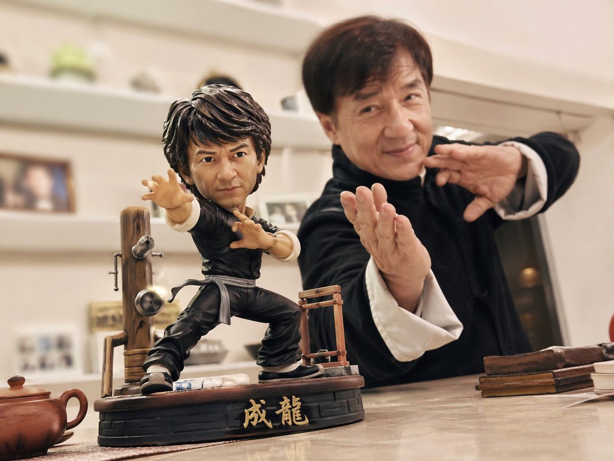 以手机随手拍摄的日常风格，桌面上摆放着一款 【成龙】【功夫】 动漫手办，动作夸张帅气，装备齐全。 同时，真实世界的对应人物也出现在镜头中，与手办摆出相似的姿势…