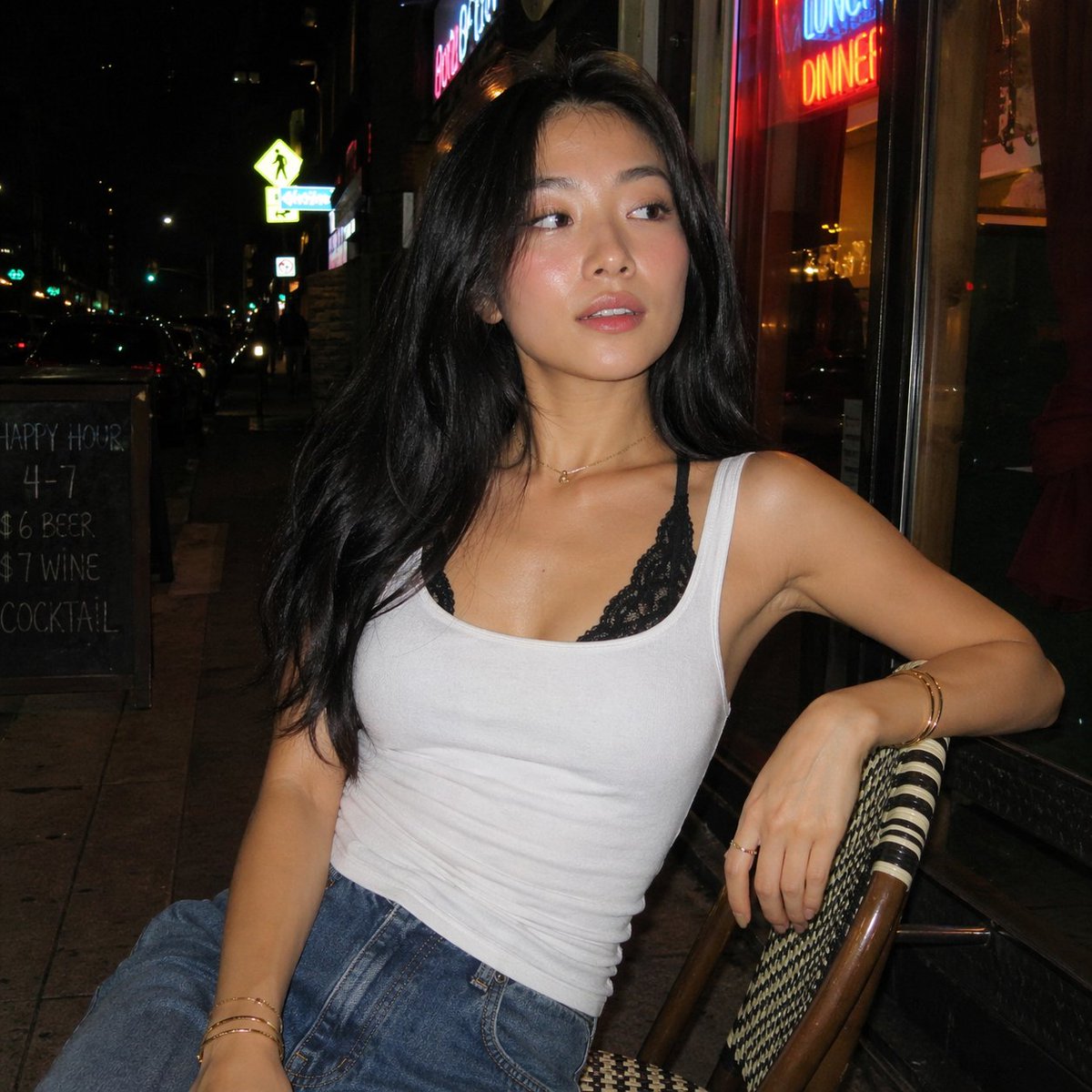 一张夜晚街头的随性人像照，照片中一位年轻亚洲女性随意地坐在一家小型城市餐厅外的藤编咖啡椅上。她有着黑色长发，妆容自然并带有柔和的光泽，身穿白色背心，内搭黑色蕾丝胸衣，下身搭配宽松…