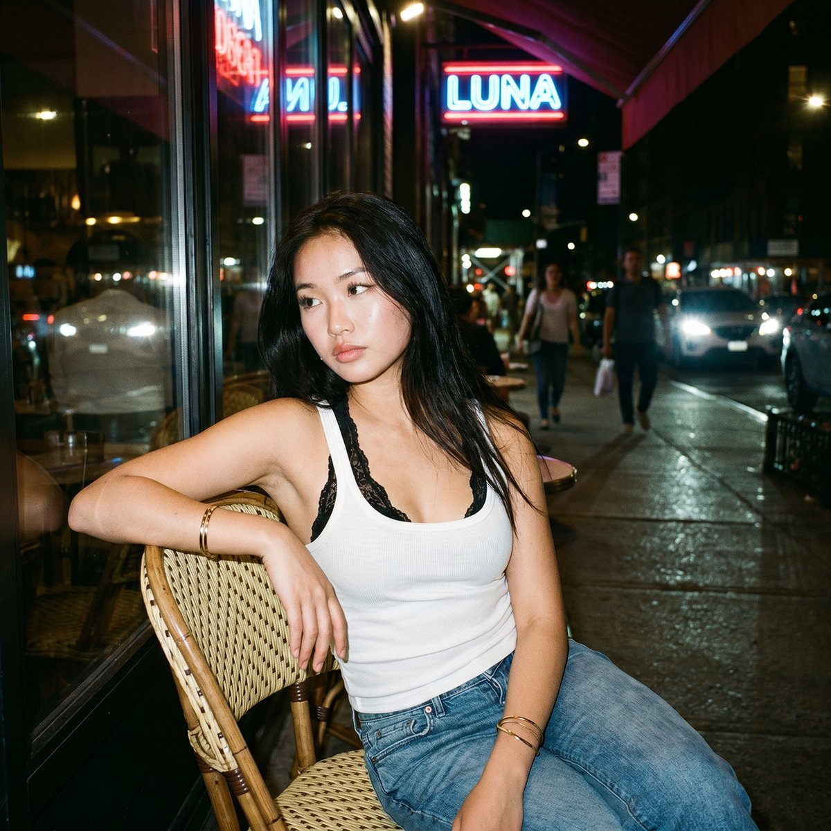一张夜晚街头的随性人像照，照片中一位年轻亚洲女性随意地坐在一家小型城市餐厅外的藤编咖啡椅上。她有着黑色长发，妆容自然并带有柔和的光泽，身穿白色背心，内搭黑色蕾丝胸衣，下身搭配宽松…