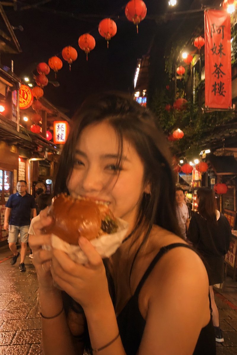 失焦的业余快照：一位亚洲超级模特站在夜晚的老台湾街道中央，发光的红灯笼照亮了整个场景。她坐在街边小吃桌前，吃着一个多汁的大汉堡，心情愉悦。暖色调。Pinterest 美学。JPG…