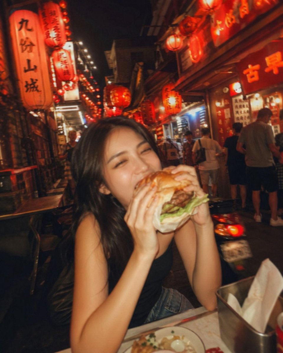 失焦的业余快照：一位亚洲超级模特站在夜晚的老台湾街道中央，发光的红灯笼照亮了整个场景。她坐在街边小吃桌前，吃着一个多汁的大汉堡，心情愉悦。暖色调。Pinterest 美学。JPG…
