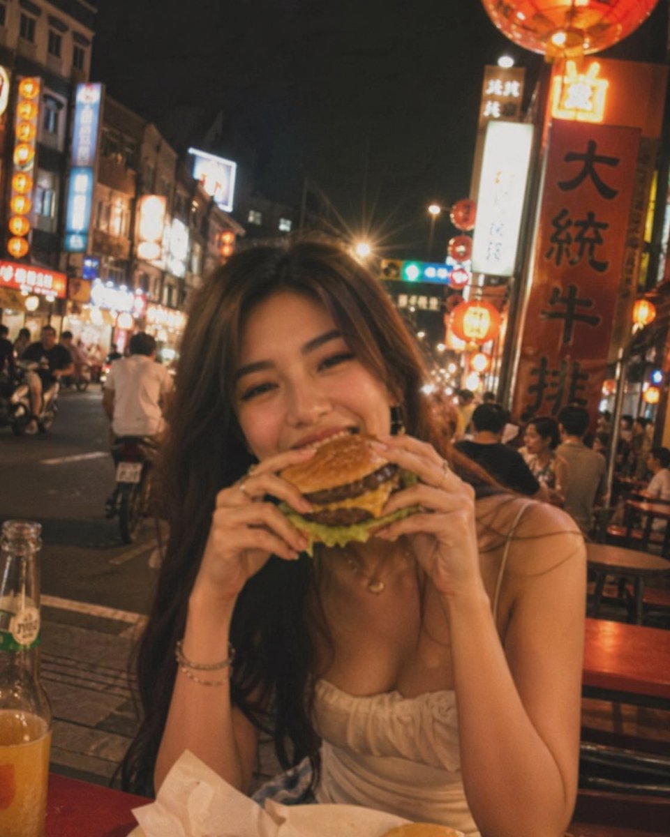 失焦的业余快照：一位亚洲超级模特站在夜晚的老台湾街道中央，发光的红灯笼照亮了整个场景。她坐在街边小吃桌前，吃着一个多汁的大汉堡，心情愉悦。暖色调。Pinterest 美学。JPG…
