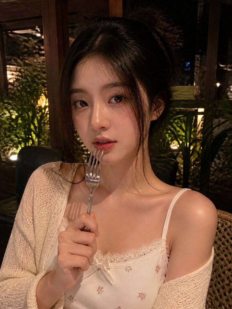 一张写实风格的弱光手机人像照片，主体是一位年轻成年女性，夜晚坐在一家温馨的咖啡馆或餐厅内。近景半身构图，画面比例约为 4:5 或接近正方形。相机举在胸口到脸部之间的高度，从近距离…