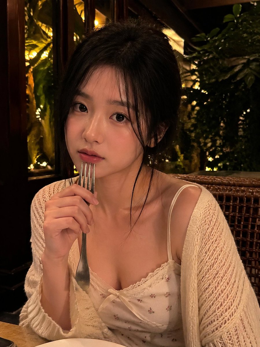 一张写实风格的弱光手机人像照片，主体是一位年轻成年女性，夜晚坐在一家温馨的咖啡馆或餐厅内。近景半身构图，画面比例约为 4:5 或接近正方形。相机举在胸口到脸部之间的高度，从近距离…