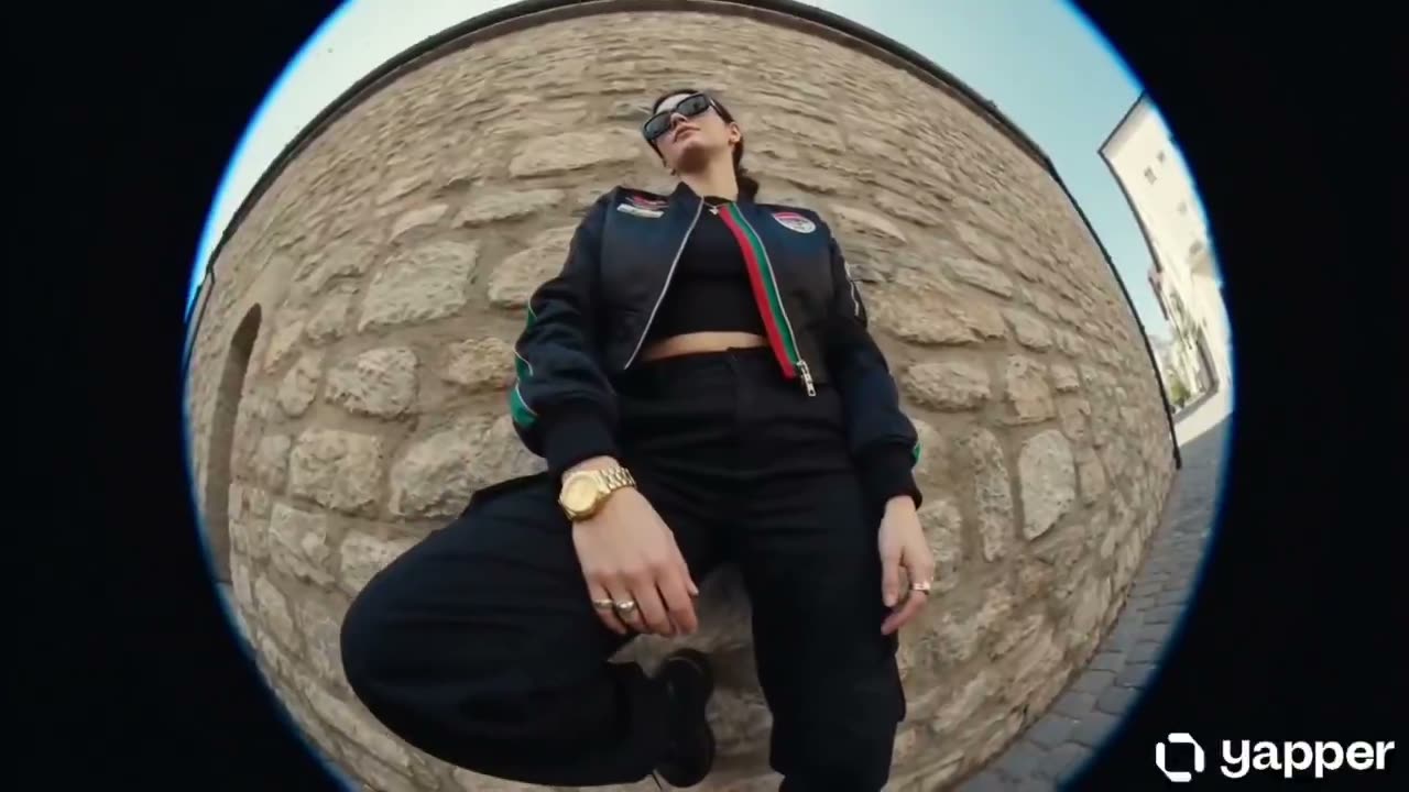 [00:00–00:01] 白天。荷兰角中景,鱼眼镜头畸变。她靠在一面素净的石墙上,一只膝盖弯曲,脚平踩在身后的墙面上。头部随着节拍轻轻上下点动,双手自然垂于…