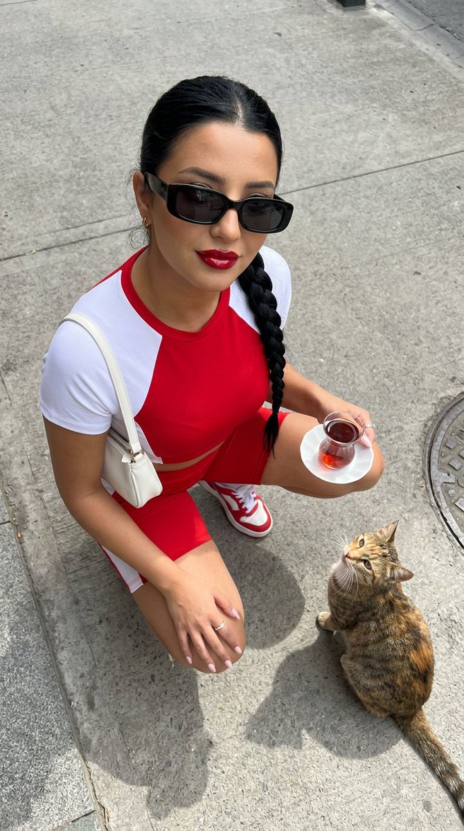 一张高角度俯视的超写实时尚摄影照片：一位时尚年轻女性站立在干净浅灰色的水泥街道上，自信而有魅力地抬头直视镜头。她留着没有头巾的深色长发，唇色为光泽的樱桃红，拥有温暖的古铜色肌肤…
