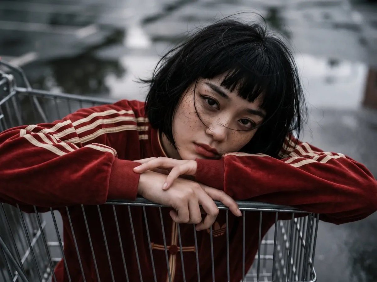 女性角色位于购物推车内部,以推车内部为视角向上望向她的脸部,戏剧性的透视畸变,一条腿靠近镜头,姿势"无所谓"且放松:,超大红色(偏红)的阿迪达斯旗袍风格天鹅绒运动套装,配米色条纹…