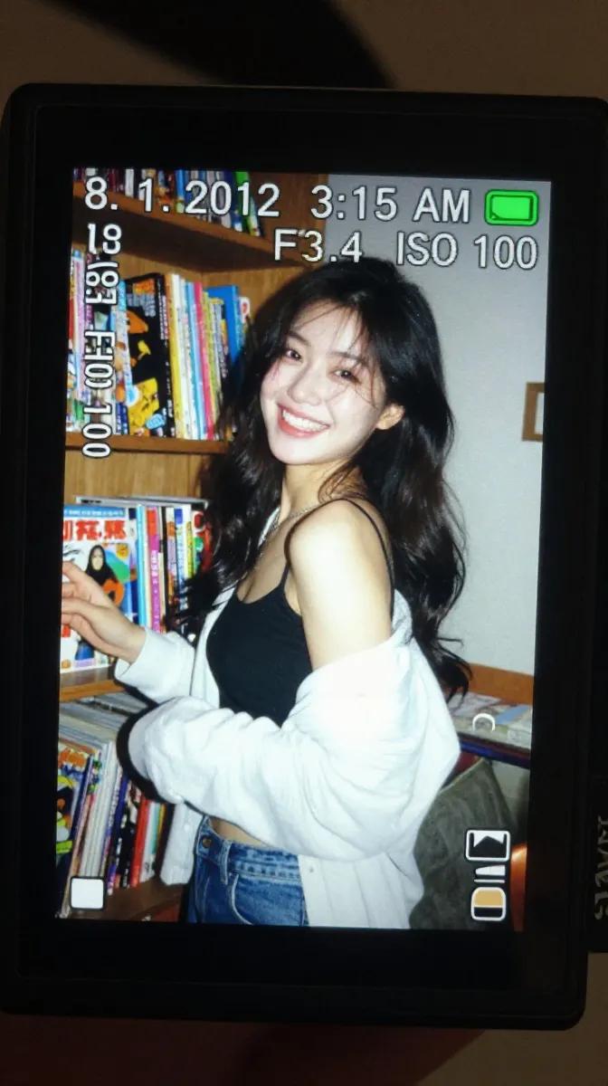 一个真实感的特写镜头，拍摄的是在昏暗室内环境中明亮发光的一台小型数码相机屏幕。LCD 屏幕上显示着一张抓拍风格的早期 2010 年代风格照片，照片中一位年轻东亚女性留着长长的深色…