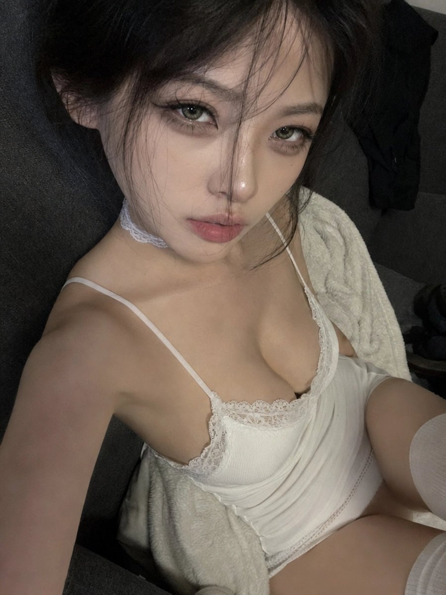 一张近距离高角度的智能手机自拍照，画面中一位年轻女子在夜晚室内斜倚着一张深灰色布艺沙发。手机举在她脸部上方，形成自然的广角畸变，头身比略微放大，上半身透视被压…