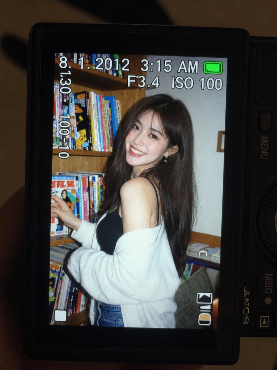 一张超精细的特写镜头：一台微型紧凑型数码相机的 LCD 屏幕在原本黑暗的室内房间中明亮发光。屏幕上是一张 2010 年代初期的随性傻瓜相机快照，画面中一位年轻东亚女性留着一头深色…