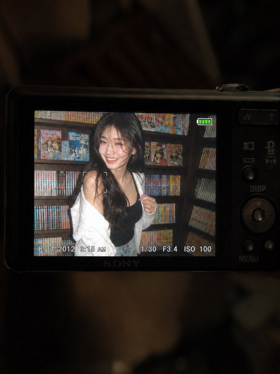 一张超精细的特写镜头：一台微型紧凑型数码相机的 LCD 屏幕在原本黑暗的室内房间中明亮发光。屏幕上是一张 2010 年代初期的随性傻瓜相机快照，画面中一位年轻东亚女性留着一头深色…