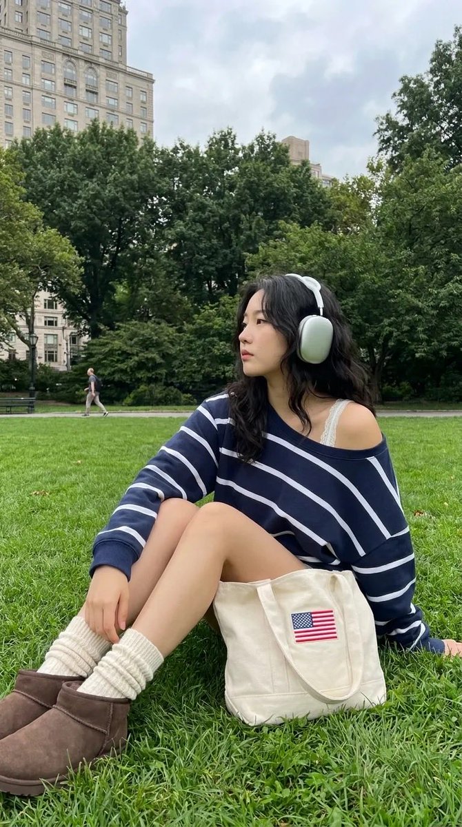 中景全身镜头:一位时髦的24岁韩国女性在像中央公园这样的知名城市公园的翠绿草坪上放松休憩。她弯曲膝盖坐着,身体微微后倾,姿态放松,同时转头看向镜头外,露出宁静沉思的侧脸。她佩戴着…