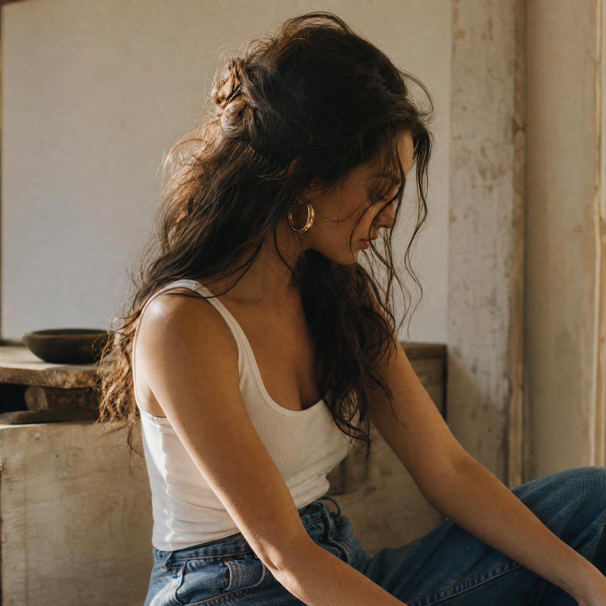 一张自然随性的中景人像照：一位女性拥有蓬松凌乱、波浪卷曲的深棕色长发，部分以半扎发样式向后别起，鬓角处有几缕碎发自然垂落，修饰着脸部轮廓。她若有所思地向下凝视…