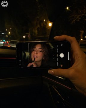 照片拍摄于夜间车内；画面中仅可见女性的一只手和一部iPhone，而年轻女性的面部仅出现在手机屏幕上。相机位于副驾驶一侧，取景框对准挡风玻璃和手机，手机由单手举…