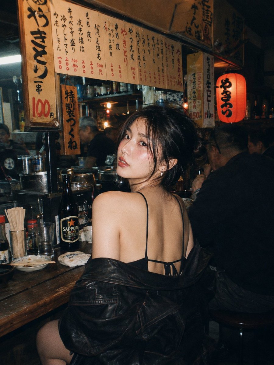 一位美丽的年轻东亚女性，留着深色波浪长卷发，妆容精致，夜晚坐在一家温馨昏暗的日本居酒屋的木质吧台前。她转过头，越过肩膀直视镜头，眼神妩媚诱人。她身穿白色背心…