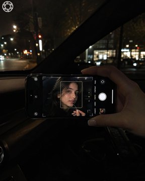 照片于夜间在车内拍摄；画面中仅露出一只女性的手和一部iPhone，年轻女性的面部仅出现在手机屏幕上。相机位于副驾驶侧，构图同时纳入挡风玻璃与手机，手机被单手直…