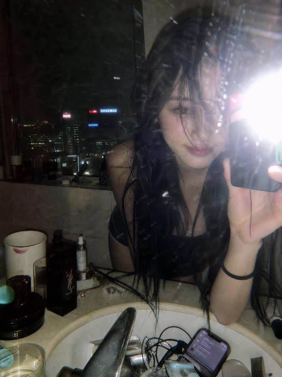 一张氛围感十足的深夜镜面自拍,拍摄于一间俯瞰璀璨城市天际线的豪华高层公寓浴室中。一位年轻女性留着略带凌乱黑色长发,身体向前倚靠在白色大理石洗手台上方,蓬松的发丝若隐若现地遮挡着一…