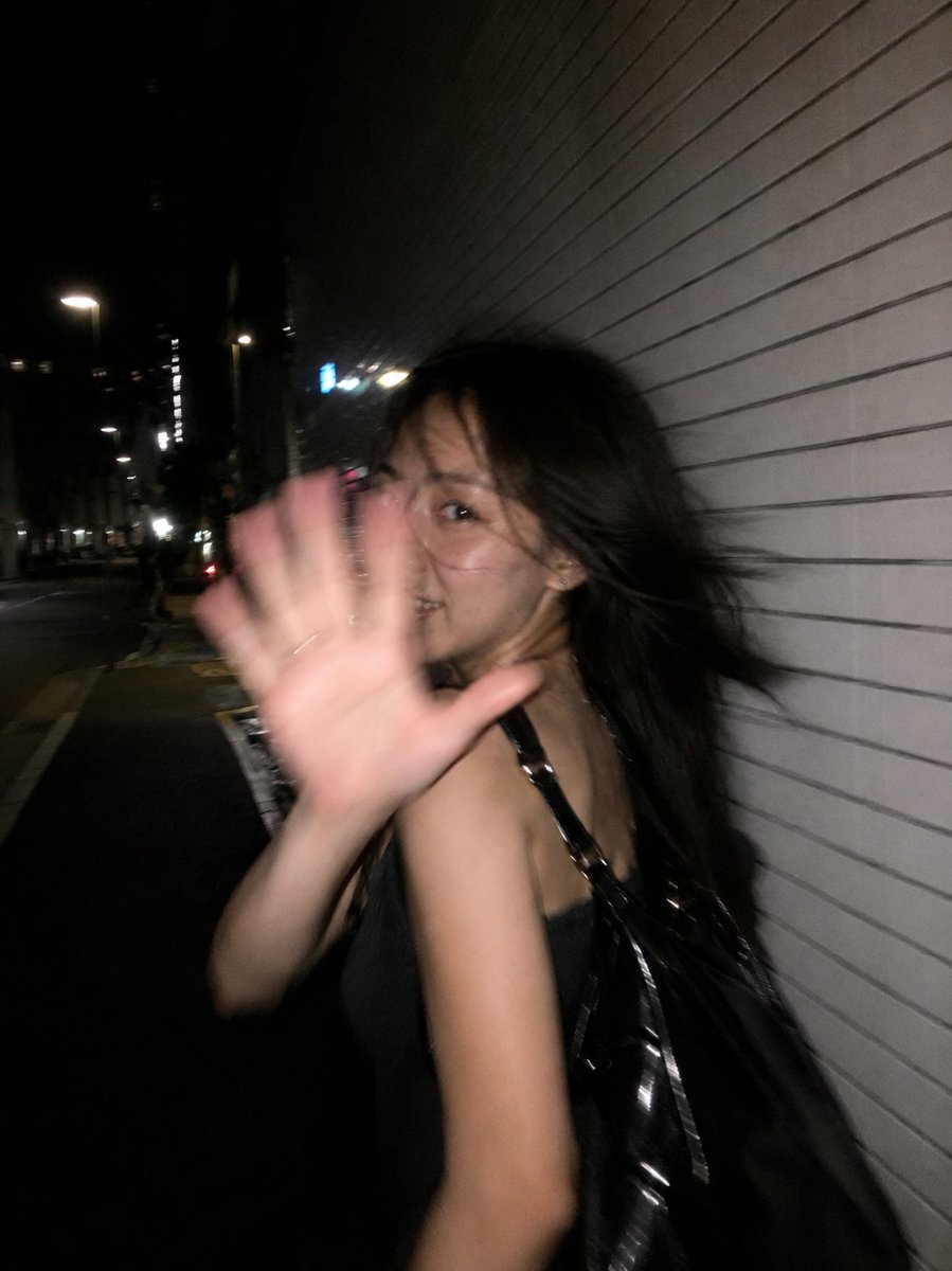超写实的抓拍式智能手机闪光灯照片,竖向构图,3:4 比例。一位年轻的东亚女性,夜晚在一段安静的城市街道上快步前行,街边是一面长长的白色瓷砖墙。以一种自发的狗仔队风格瞬间捕捉——她…