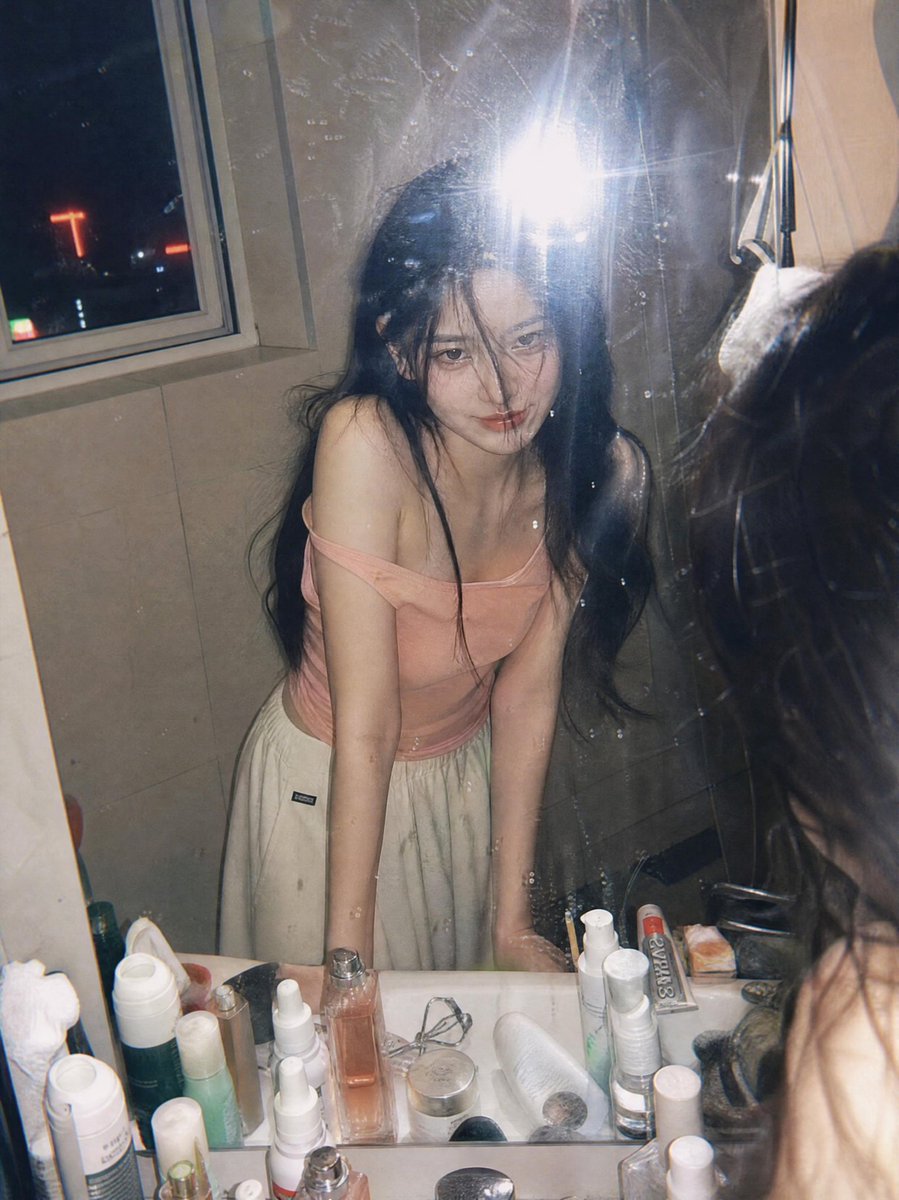 超写实抓拍胶片摄影人像：一位年轻女子夜晚俯身在浴室洗手台前，透过镜子凝视自己。画面通过一面略有污渍的镜子拍摄，呈现自然的反射与细腻的瑕疵。她留着黑色长发，部分发丝遮住面庞，皮肤柔…
