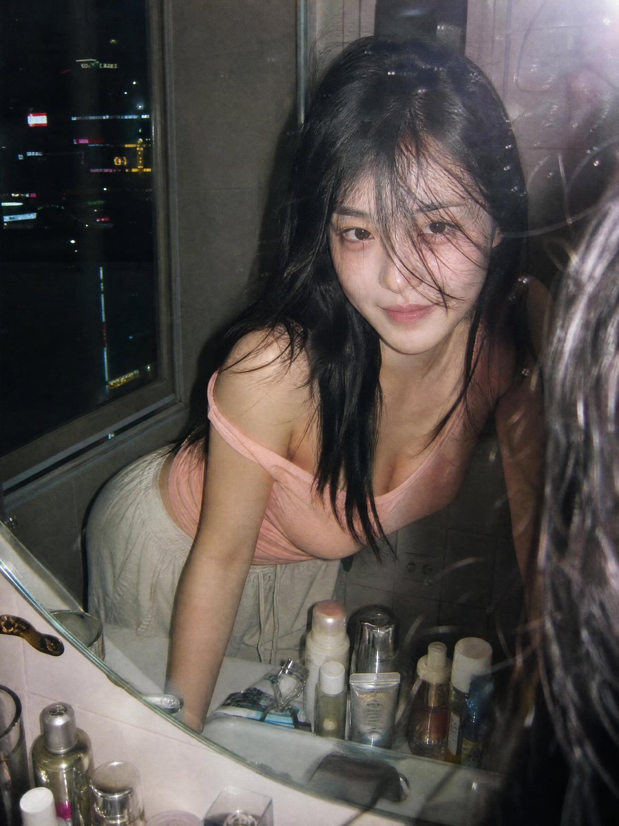 超写实抓拍胶片摄影人像：一位年轻女子夜晚俯身在浴室洗手台前，透过镜子凝视自己。画面通过一面略有污渍的镜子拍摄，呈现自然的反射与细腻的瑕疵。她留着黑色长发，部分发丝遮住面庞，皮肤柔…