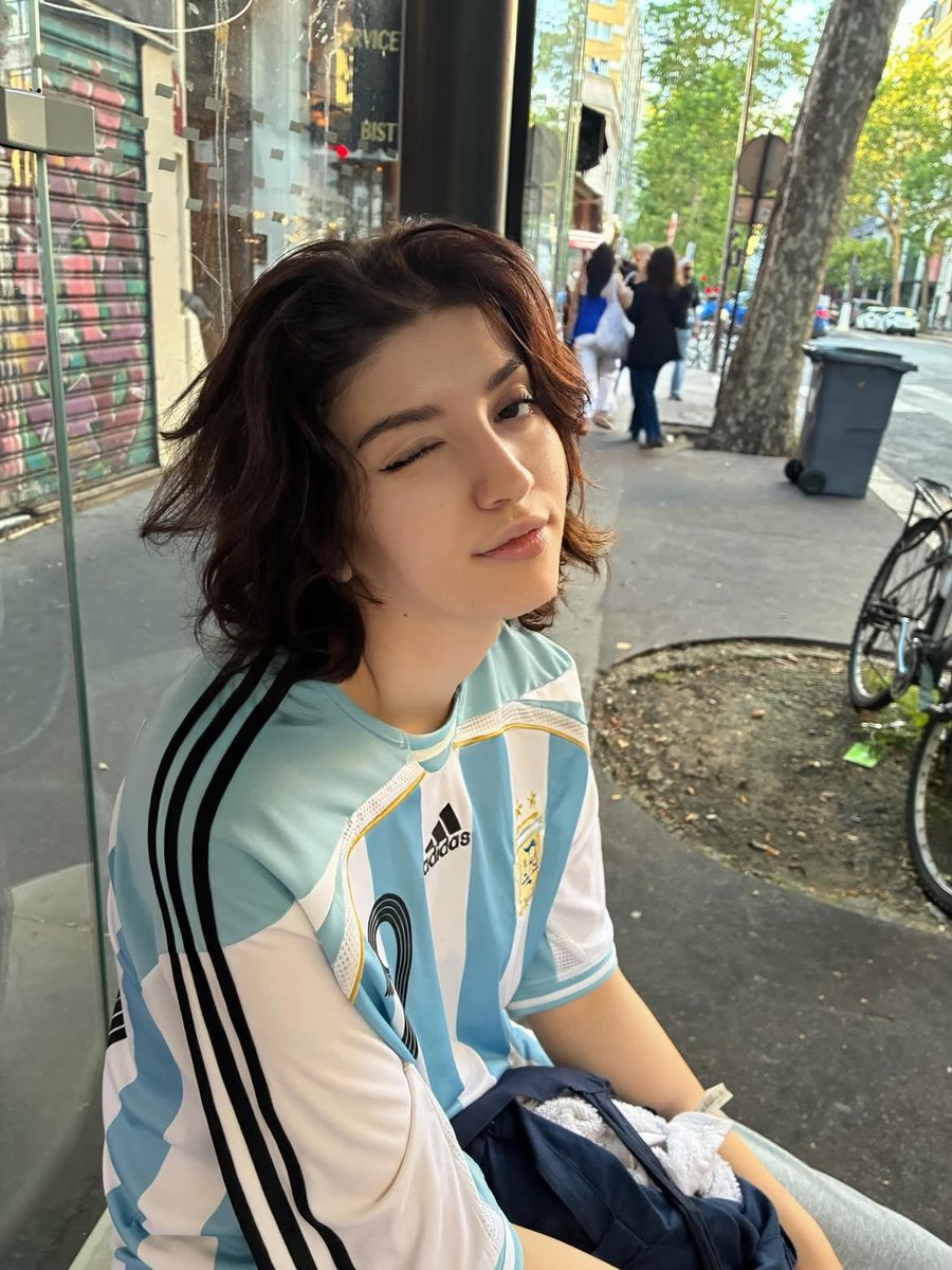 超写实的随性人像摄影：一位留着短波浪深色头发的年轻人，身着阿根廷国家足球队宽大球衣（19号），在黄金时段坐在城市公交车内和公交车站上。柔和温暖的阳光透过大窗户洒进来，在脸上形成戏…