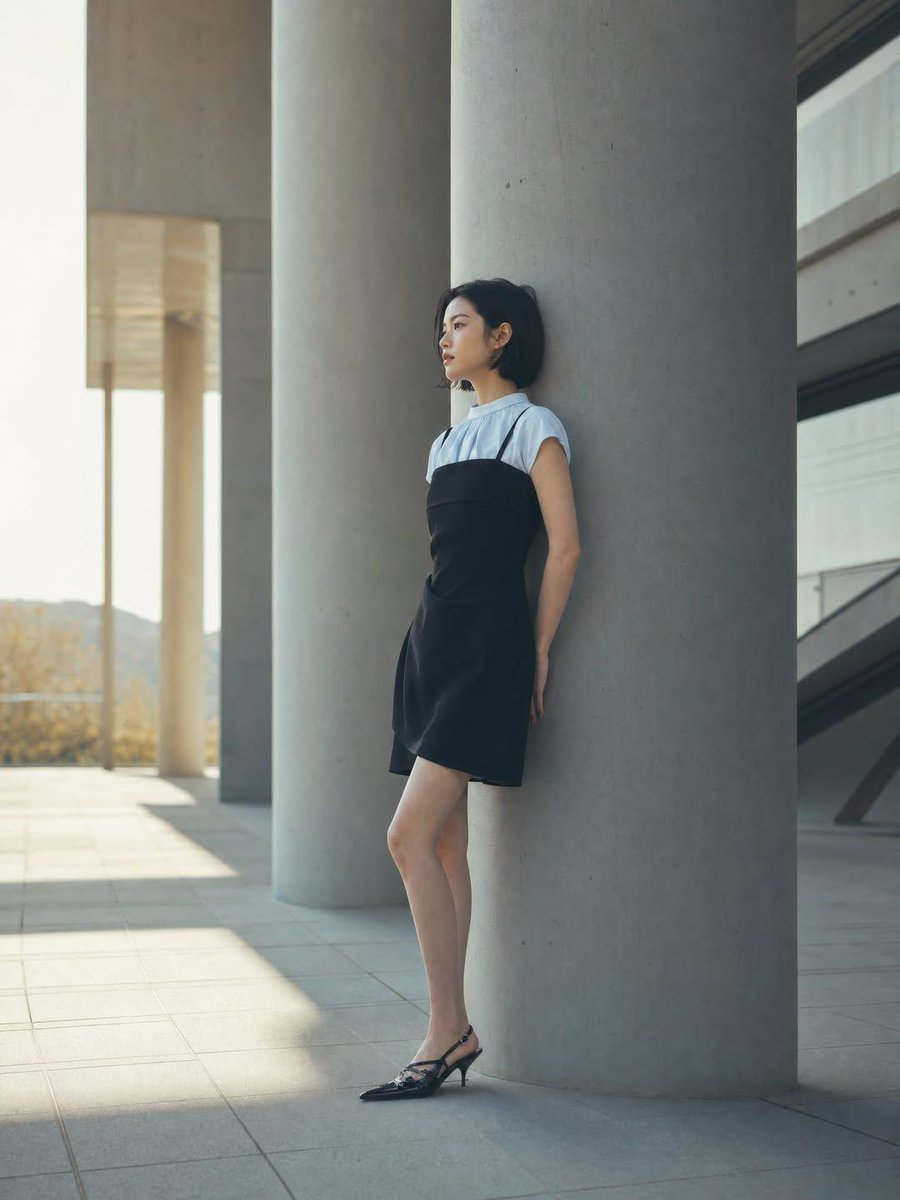 一位身材纤细的年轻女性，留着黑色短波波头发型，靠在现代建筑广场上一根巨大的混凝土柱上，身穿黑色吊带裙内搭白色短袖上衣，脚踩黑色尖头后跟凉鞋，妆容自然柔和，侧脸若有所思，极简时尚大…