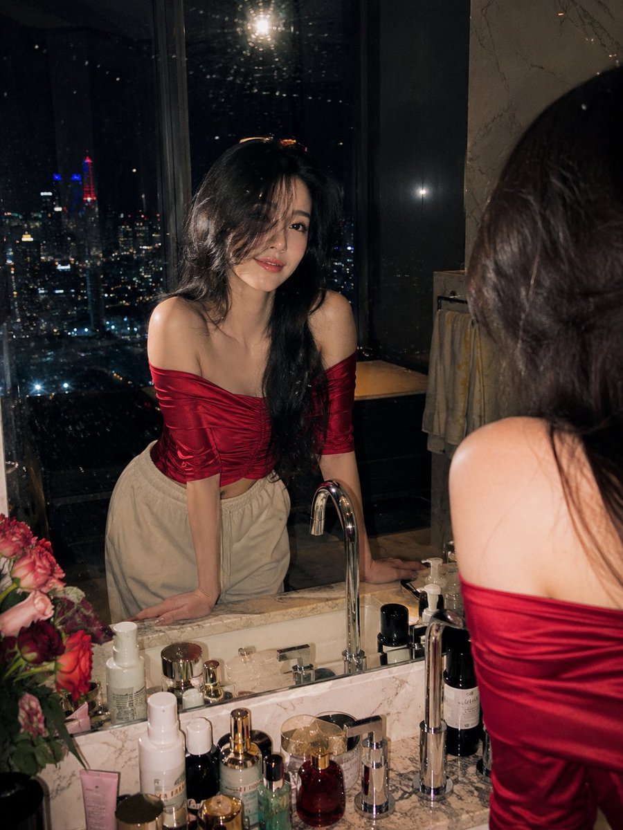 超写实电影感夜景人像摄影:一位美丽的年轻女子站在顶层豪宅豪华大理石浴室梳妆台前,俯瞰夜晚璀璨的城市天际线。她身穿酒红色光泽缎面露肩上衣,面料垂坠优雅,搭配宽松米白色休闲裤。深色长…