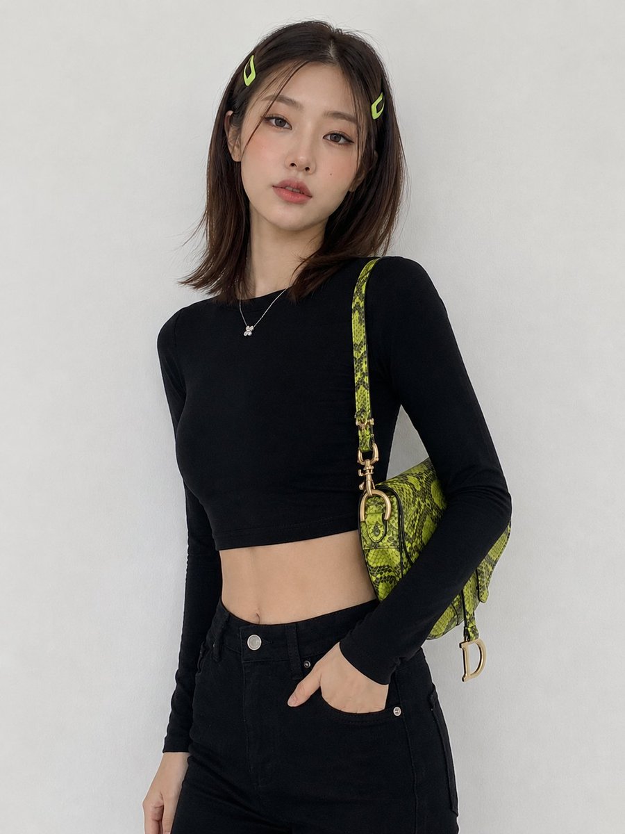 创建一幅图像：一位年轻韩国女性，中长棕色头发，两侧别着青柠绿色发夹，倚靠在一面纯白色墙壁前摆姿势，身穿黑色短款长袖上衣、黑色高腰长裤，肩上斜挎一款色彩鲜艳的青…