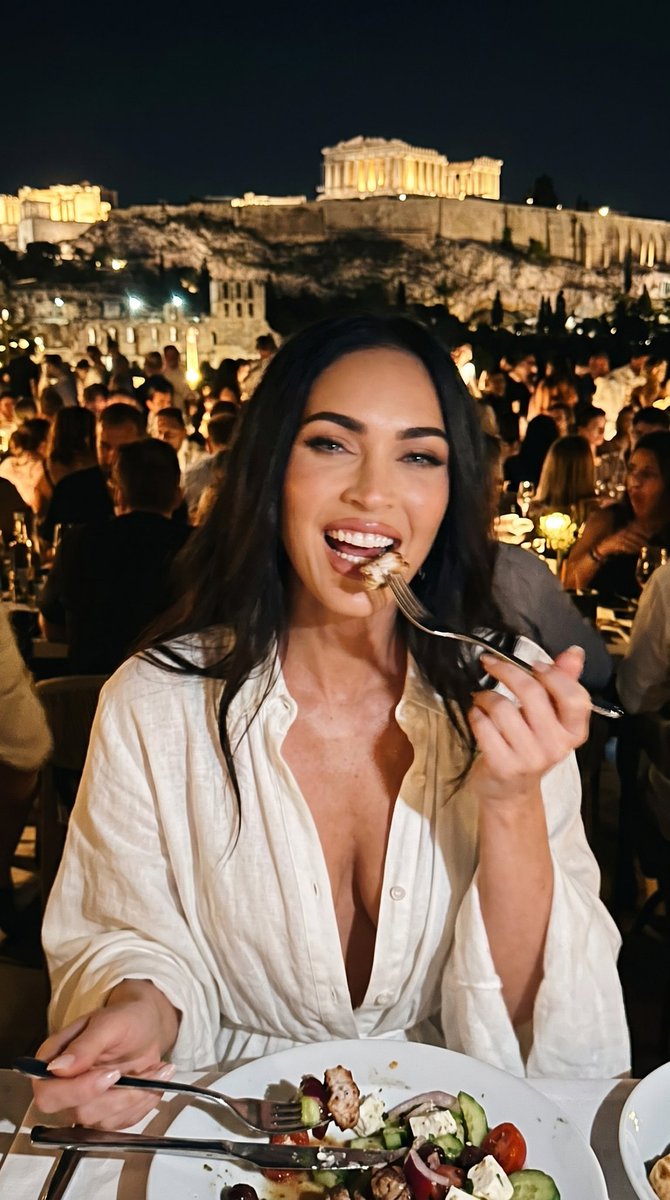 一张微妙模糊、低画质的特写业余 iPhone 照片，内容是一位惊艳的年轻 Megan Fox（梅根·福克斯），在雅典帕特农神庙附近一家高档拥挤的餐厅里，身穿一…
