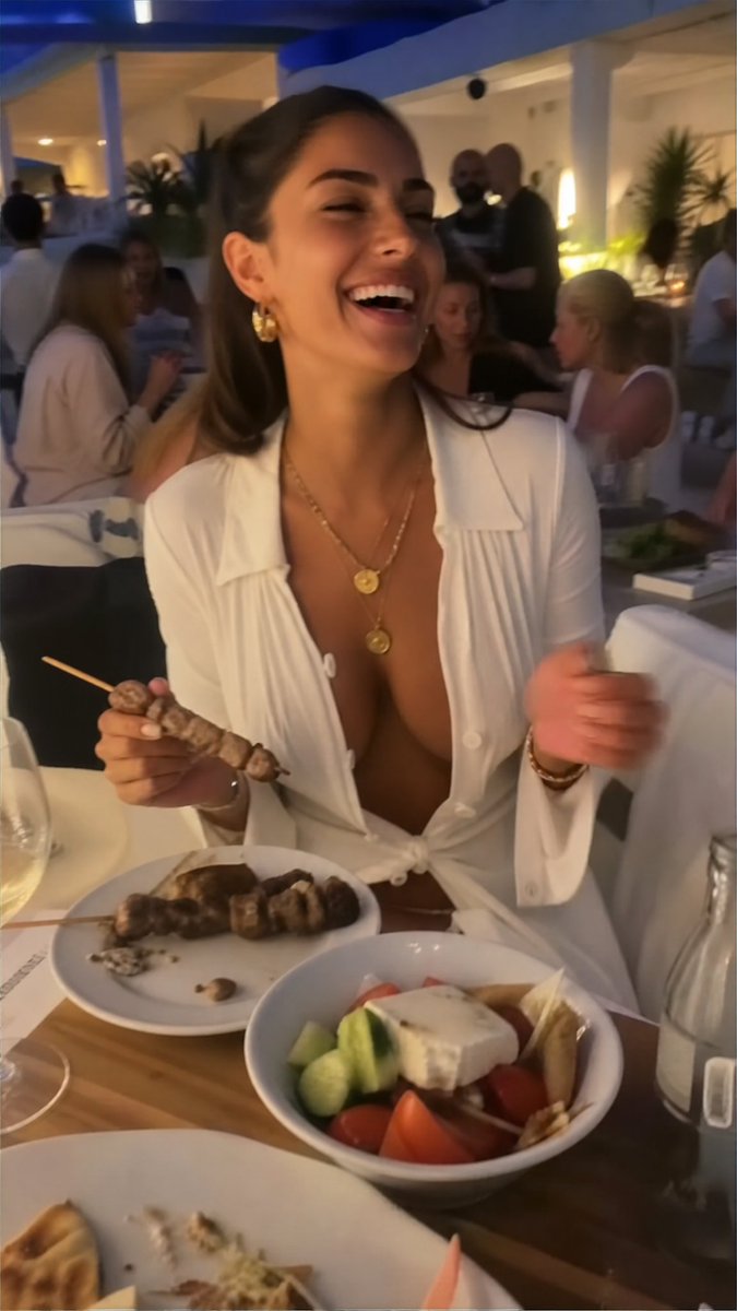 一张微妙模糊、低画质的特写业余 iPhone 照片,内容是一位惊艳的年轻 Megan Fox(梅根·福克斯),在雅典帕特农神庙附近一家高档拥挤的餐厅里,身穿一袭全白色女神式未扣纽…