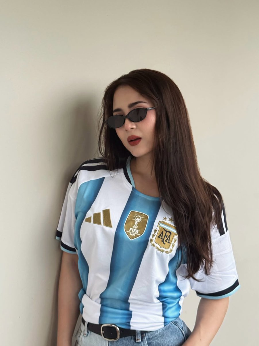 超现实风格的时尚人像摄影：一位时髦的年轻女性，身穿不同国家队的足球球衣（阿根廷、葡萄牙、巴西），在室内一面干净极简的米白色墙壁前自信地摆姿势。佩戴时髦的窄框黑色太阳镜，肌肤白皙无…