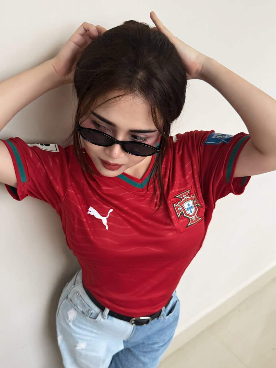 超现实风格的时尚人像摄影：一位时髦的年轻女性，身穿不同国家队的足球球衣（阿根廷、葡萄牙、巴西），在室内一面干净极简的米白色墙壁前自信地摆姿势。佩戴时髦的窄框黑色太阳镜，肌肤白皙无…