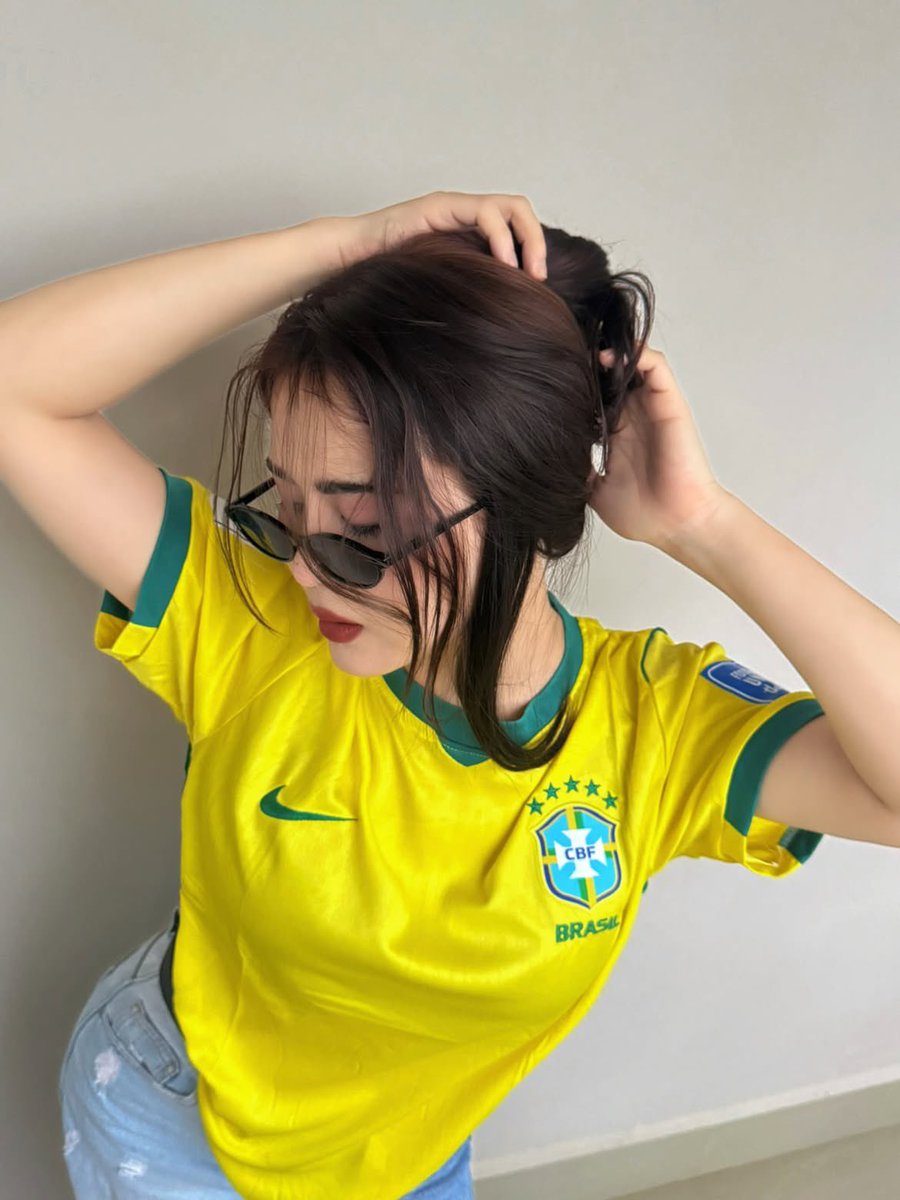 超现实风格的时尚人像摄影：一位时髦的年轻女性，身穿不同国家队的足球球衣（阿根廷、葡萄牙、巴西），在室内一面干净极简的米白色墙壁前自信地摆姿势。佩戴时髦的窄框黑色太阳镜，肌肤白皙无…