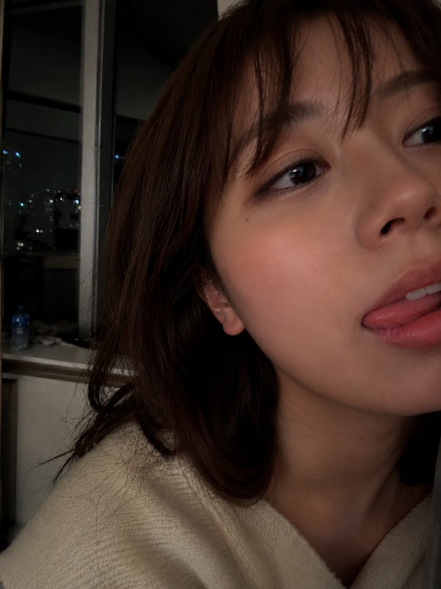 室内夜景中，使用智能手机摄像头拍摄的年轻女性超写实特写自拍肖像。肌肤柔和自然，肤质光滑细腻，双颊微微泛红，嘴唇粉嫩水润，表情俏皮，舌头轻触上唇。一双深色的大眼睛略带灵动，微微侧向…