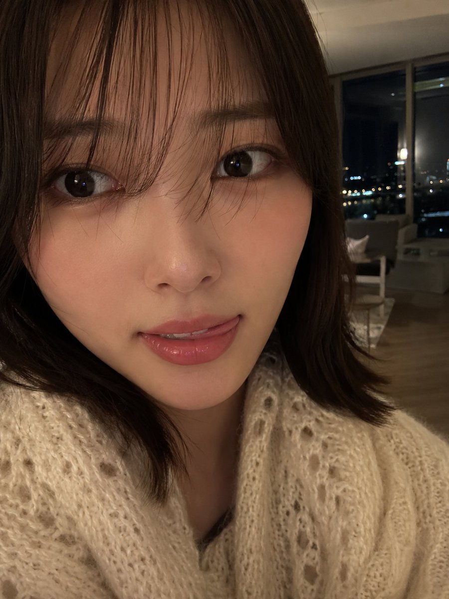 室内夜景中，使用智能手机摄像头拍摄的年轻女性超写实特写自拍肖像。肌肤柔和自然，肤质光滑细腻，双颊微微泛红，嘴唇粉嫩水润，表情俏皮，舌头轻触上唇。一双深色的大眼睛略带灵动，微微侧向…