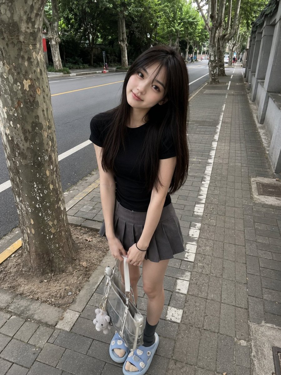 年轻的日本女孩，长直深棕色头发配柔和刘海，身着修身黑色T恤、灰色百褶迷你裙、灰色袜子与浅蓝色拖鞋，随意站在绿树成行的安静道路旁的人行道上。微微前倾，露出可爱自然的微笑，手里拿着一…