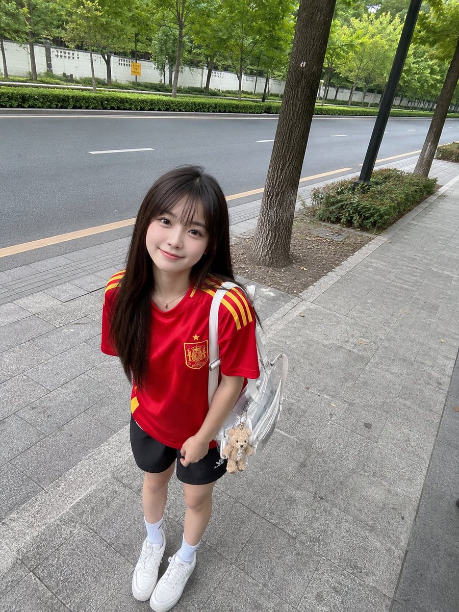 年轻的日本女孩,留着长直的深棕色头发和柔软的刘海,身穿红色西班牙国家足球队球衣、黑色运动短裤、白色中筒袜和白色运动鞋,悠闲地站在绿树成荫的安静道路旁的人行道上。她微微前倾,露出可…
