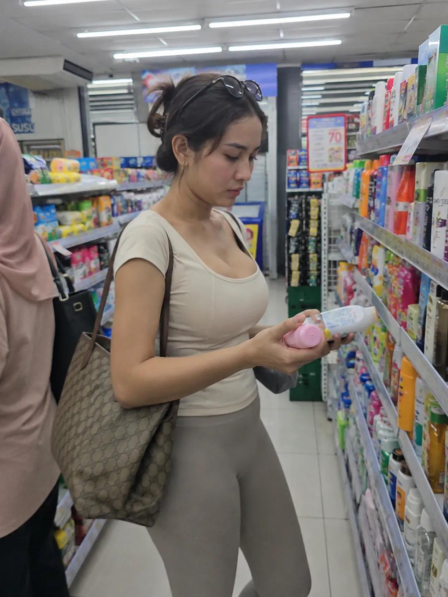摄影级写实的随性生活照片：一位年轻女性在光线明亮的便利店过道中购物。侧面视角，中全景全身镜头。她站在两排货架之间，货架上摆满了色彩缤纷的个人护理和家居用品；她一手正仔细阅读一个小…