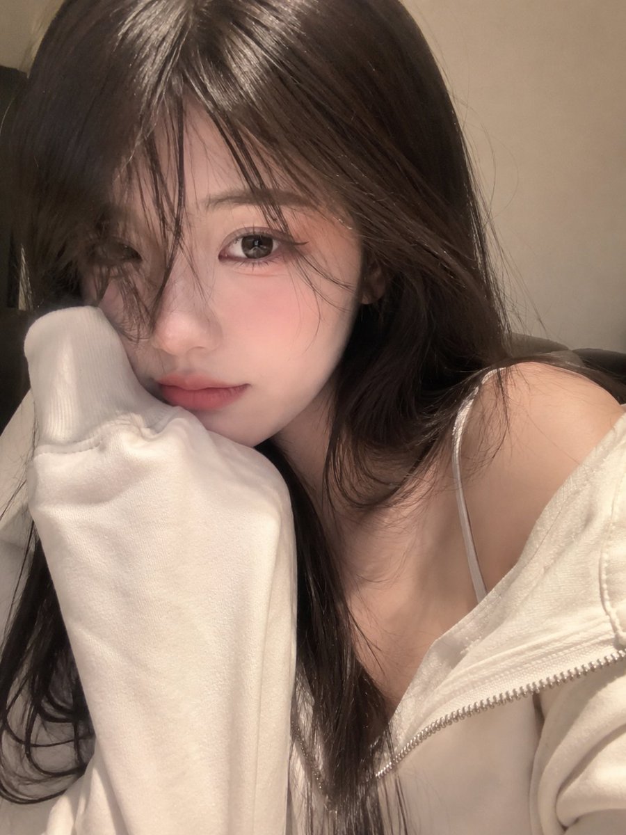 一张照片般逼真的特写自拍,一位可爱的年轻女性,脸部占据画面的大部分,头部微微倾斜,脸颊舒适地靠在一件宽松的奶油色连帽衫袖子里。柔顺的长发配有柔和的八字刘海遮住一只眼睛,大而有神的…