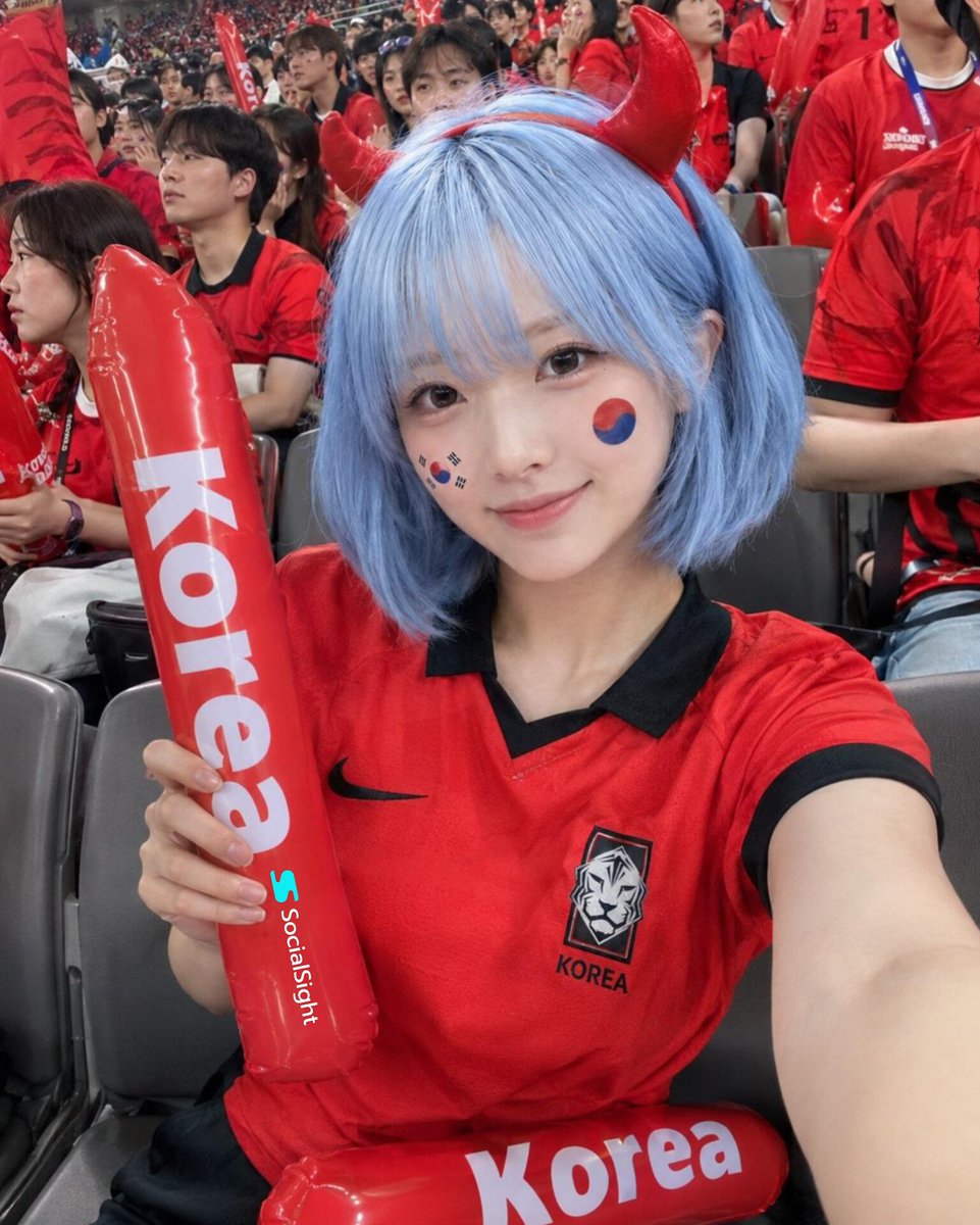 在一座座无虚席的体育场内，国际足球比赛期间，一位美丽的足球迷坐在看台上拍摄的极致写实自拍，齐耳短款粉蓝色波波头，闪亮的红色恶魔角发箍，柔和自然妆容，明亮有神的眼睛，对着镜头露出淡…