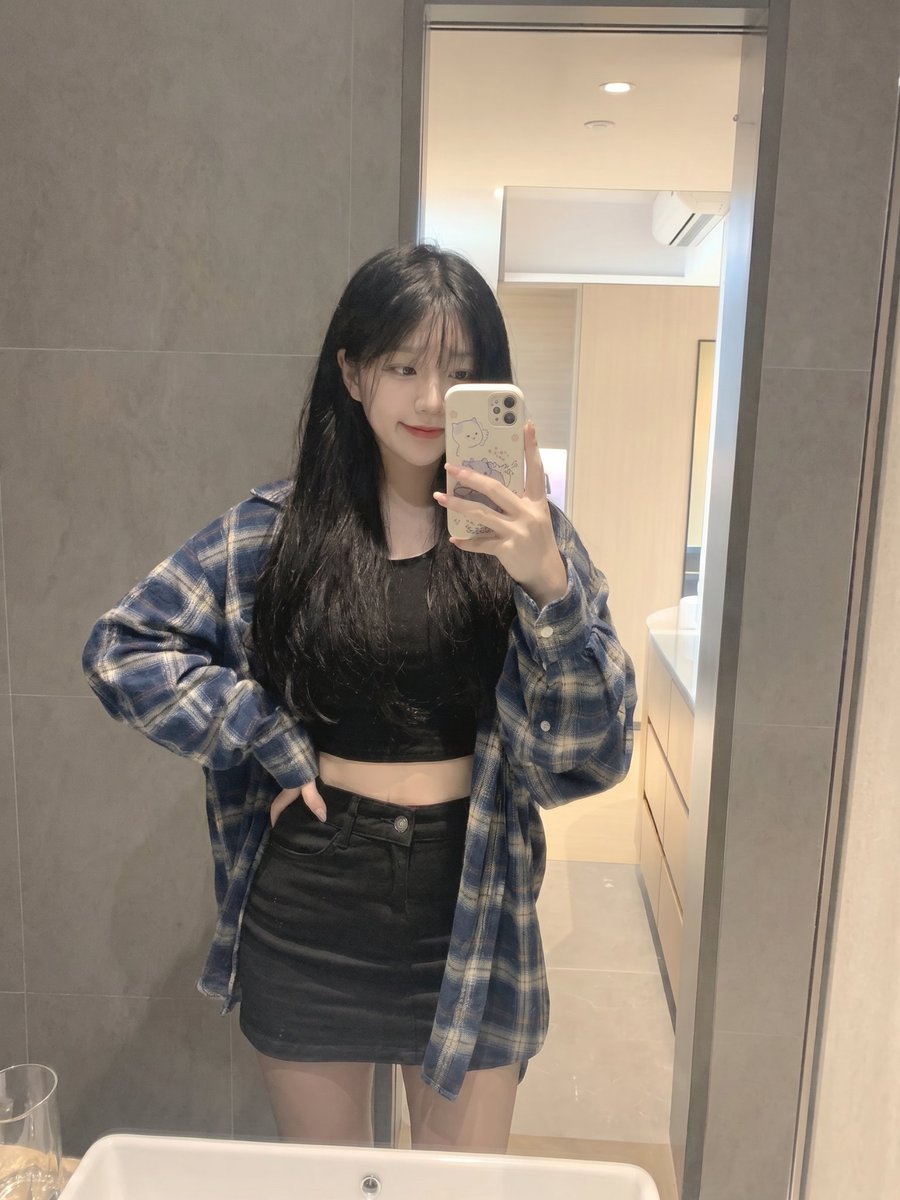 留着长长的黑色直发和柔软稀疏刘海的年轻日本女孩,内穿黑色短款露脐上衣,外穿 oversized 蓝色格纹法兰绒衬衫,搭配黑色迷你短裙。站在浴室镜子前,拿着配有可爱卡通保护壳的智能…