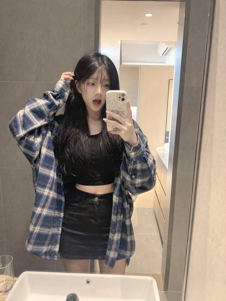 留着长长的黑色直发和柔软稀疏刘海的年轻日本女孩,内穿黑色短款露脐上衣,外穿 oversized 蓝色格纹法兰绒衬衫,搭配黑色迷你短裙。站在浴室镜子前,拿着配有可爱卡通保护壳的智能…
